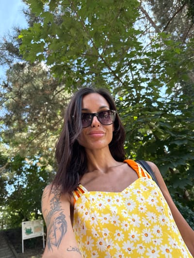 A woman is captured in a selfie, standing outdoors with lush green trees and a bright blue sky in the background. She is wearing sunglasses and a yellow dress adorned with a pattern of white daisies. Her left arm is visible, displaying several tattoos. The image appears to have been taken in Grivitsa, Bulgaria, possibly in a park or natural setting.