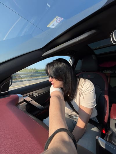 An indoor shot captures two individuals inside a car, with the focus on a woman in the passenger seat and the arm of the photographer in the foreground. The woman, with long dark hair and sunglasses, is looking out the side window. The interior of the car features a sunroof, a red leather-accented dashboard, and car seats with red and black upholstery. Outside the car, a highway with a metal barrier and vegetation is visible. The scene is illuminated by natural light, suggesting it is daytime. The image does not provide any specific information or context to determine a location in the city of Фёдорово, Russia.
