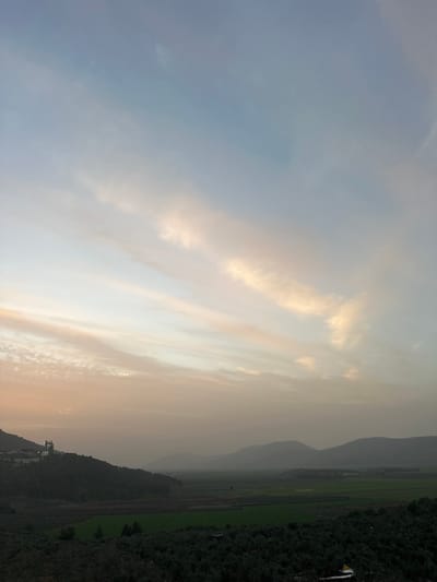 The image displays a serene landscape scene, likely captured during either sunrise or sunset, given the warm and soft lighting conditions. The primary subject is the sky, which occupies a significant portion of the frame and exhibits a beautiful gradient of colors. On the left side of the sky, the colors are a soft blue-grey, transitioning smoothly towards the right into warmer hues of golden yellow and light orange, suggesting the sun is either rising or setting in that direction. Streaks of wispy, elongated clouds are visible across the sky, particularly catching the golden light on the right, creating soft highlights and shadows.
Below the expansive sky, a layered landscape unfolds. In the mid-ground and background, a series of undulating mountains or hills stretch across the horizon. These mountains appear somewhat hazy and muted in color, indicating distance and atmospheric perspective, a common visual effect during dawn or dusk.
To the right side of the frame, a dark, densely vegetated or forested hill slopes upwards, contrasting with the lighter sky and distant mountains. This hill appears closer to the viewer. Adjacent to this hill and extending towards the center-left, there are expansive, flat green fields, likely agricultural land such as rice paddies, stretching into the distance. These fields appear verdant and well-tended.
A small, indistinct white object, possibly a flag or a small structure, can be seen on the upper part of the dark, forested hill on the far right. The overall impression is one of tranquility and natural beauty, highlighting the transition of day with soft light and atmospheric depth over a rural, mountainous region. No discernible text is visible in the image.