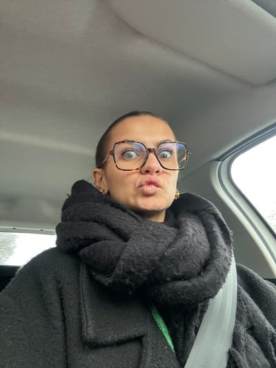 A female individual is depicted within the interior of a vehicle, located in Saint-Cyprien, France. The subject wears large, square, tortoiseshell-patterned eyeglasses, a black, textured coat, and a thick, black knitted scarf wrapped around her neck. Two gold-colored stud earrings, appearing possibly floral or star-shaped, are visible on her earlobes. A grey seatbelt is secured across her chest. Her hair is pulled back, and her face exhibits wide eyes and pursed lips, forming a "kissing" or "fish-face" expression, indicative of a posed self-portrait. A portion of a bright green garment is visible beneath the scarf and coat on the left side. The vehicle's interior features a light grey headliner. The exterior environment visible through the right side window is indistinct, suggesting an overcast sky.