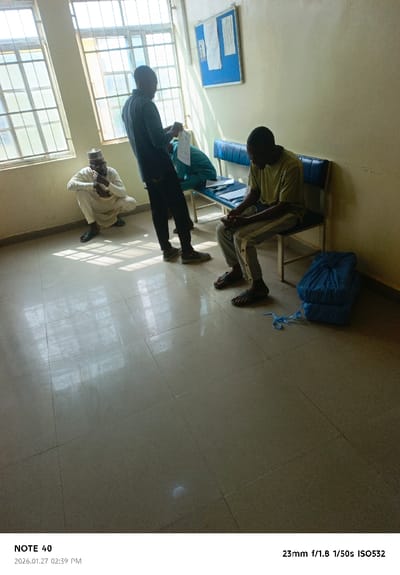The image depicts an interior scene in Gwammaja, Nigeria, likely a waiting area or office. The room features light-colored tiled flooring and plain, light-colored walls. Two large windows with grid patterns on the left wall allow natural light to enter, casting distinct shadows on the floor. A blue-framed bulletin board with several documents is affixed to the right wall, above a long blue bench with a metal frame.

Four adult males are present. On the far left, an individual with a light brown beard and a cap, wearing light-colored traditional attire, is crouched with an elbow resting on a knee. In the center, an individual in dark long-sleeved clothing and trousers stands, examining a white paper document held in both hands. Behind him, an individual in light blue clothing is seated on the blue bench; a document is visible on the bench beside this person. To the right, an individual in a light-colored short-sleeved shirt and light trousers with side stripes is seated on the blue bench, looking downwards with hands clasped. On the floor to the right of the seated individual, two neatly stacked bundles of blue fabric or material, tied with light blue ribbon, are visible.