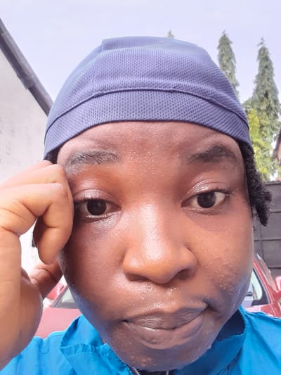 A close-up shot shows a person with dark skin, possibly male, wearing a blue head covering and a blue shirt. The skin appears slightly wet, possibly due to sweat. The person's face is turned slightly to the side, with one hand partially visible near the chin. The background is blurry, but suggests an outdoor environment with foliage and a red vehicle. There is no clear indication that this is specifically in the city of Umuigbekwo, Nigeria.
