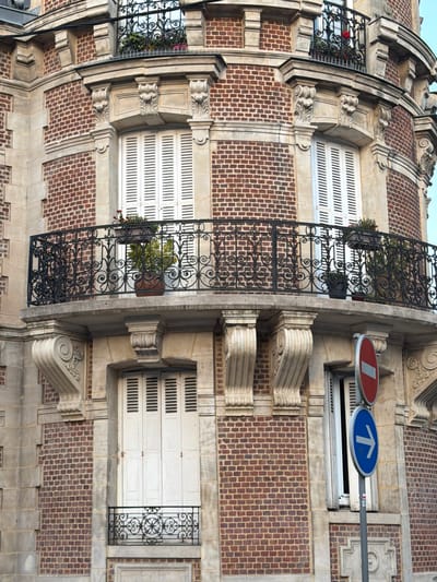 This image shows a close-up of the facade of a building in Dieppe, France, featuring a rounded corner. The architecture is characterized by red brick walls and light-colored stone accents around windows and architectural features. A wrought-iron balcony with intricate scrollwork spans across the curved section of the building. Two windows with white slatted shutters are visible on the upper level, with potted plants on the balcony in front of them. Below the balcony, another window with similar shutters and a smaller, decorative wrought-iron railing is present. To the right of the facade, a street sign pole is visible, holding a red and white "no entry" sign stacked above a blue circular sign with a white arrow pointing to the right. The sky is a pale blue.