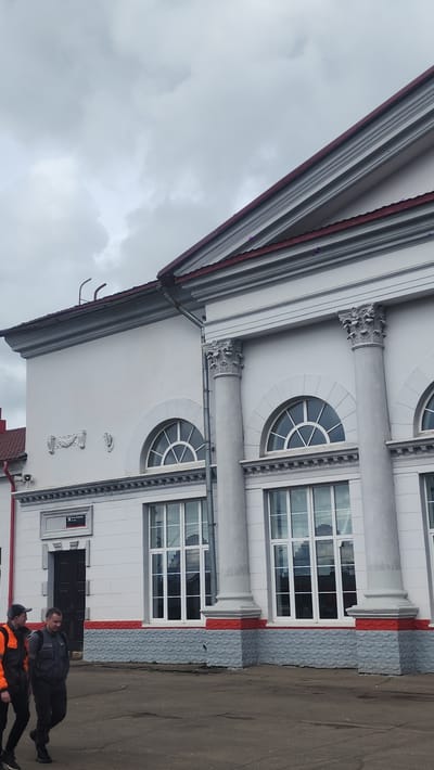 The image shows the exterior of a white building with classical architectural elements, including columns and arched windows. The building is located in Izdeshkovo, Russia. Two individuals are present in the foreground near an entrance, one wearing an orange vest. The sky is overcast.