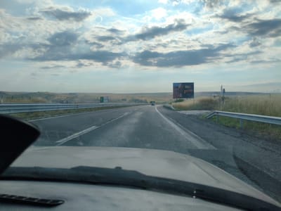The image captures a view from inside a vehicle driving on a highway. The sky is partly cloudy with patches of blue, and the sun is visible, casting rays through the clouds. A billboard displaying an advertisement for "Nuii" and possibly an ice cream product is positioned to the right of the highway. The highway itself is asphalt, with visible lane markings. Guardrails line the sides of the road. The surrounding landscape appears rural, with dry, grassy hills and fields stretching towards the horizon. There are a few scattered trees and some distant structures. No people are directly visible within the frame, other than the implied driver of the vehicle from which the photo is taken. The overall impression is that of a journey along a country road on a partly cloudy day. There is no indication of the city of Vratitsa, Bulgaria in the image.
