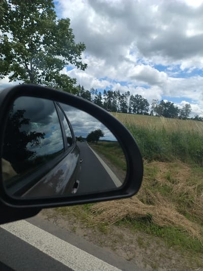 The image shows a view from a car driving on a road in Olešná, Czechia. The driver's side rearview mirror reflects a dark car and the surrounding landscape. The road has a white line marking the edge. To the right of the road is a field with dry grass and green vegetation. A line of trees is visible in the background, and the sky is partly cloudy with some blue sky showing through.