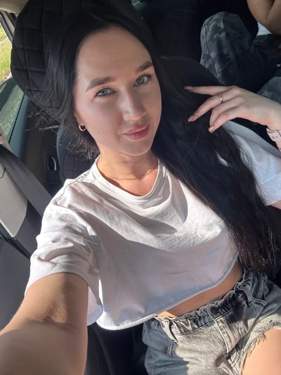 A young woman with long, dark hair and blue eyes is taking a selfie inside a car. She is wearing a white crop top and denim shorts. She has a gold necklace and a ring on her finger. The car interior is visible, including a seatbelt and a headrest. The scene is illuminated by sunlight.
