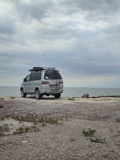 An overcast day at the coast of Aqshuqyr, Kazakhstan. A silver minivan is parked on a rocky, shell-strewn beach near the water's edge. A person in light blue shorts and a tan shirt is kneeling on the ground facing away from the camera, with their head bowed towards the earth. The sea is calm and stretches to the horizon. Birds are visible in the sky.