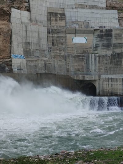 This media file depicts a section of a large concrete dam with water cascading over its edge and churning in the river below. The dam's surface is weathered and marked with graffiti. Some of the visible text in the graffiti appears to be in Cyrillic script. A large opening or tunnel is visible within the dam structure, from which water is also flowing, contributing to the torrent. The surrounding landscape suggests a rocky, mountainous terrain, with a brown, rocky cliff face visible to the left of the dam. The water in the river is a murky green, with white foam and turbulence indicating a strong current and significant flow. The overall scene conveys a sense of raw natural power controlled by human engineering. The text elements, though varied in their legibility, suggest that this is a place with some local history or at least a history of human presence and expression. The weather appears to be overcast or generally subdued, with no strong sunlight casting sharp shadows. The time of day is not clearly discernible.