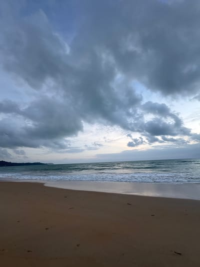 The image captures a serene beach scene in Ban Bang Niang, Thailand, under a cloudy sky. The foreground is dominated by a sandy beach with visible footprints and a few scattered dark debris, likely twigs or seaweed. The sand transitions to the sea, where gentle waves with white foam are breaking on the shore. The ocean water is a deep teal color. In the distance, a lush green shoreline with trees is visible, stretching across the horizon. The sky above is filled with dramatic, dark gray clouds, interspersed with patches of lighter gray and hints of blue sky peeking through. The overall lighting suggests it is either early morning or late afternoon, with a soft, diffused light casting subtle shadows.
