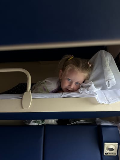 A young Caucasian girl with blonde hair in two pigtails is lying on her side on a bunk bed in what appears to be a train compartment. She is looking at the camera with wide blue eyes. She is wearing a patterned pajama set and is partially covered by a white sheet. A yellow hair tie is visible in her hair. The bunk bed has a beige mattress and a white rail. Below the bunk, blue seating is visible, along with some bags and a white electrical outlet with buttons. The background is dark, suggesting the lighting is dim. The image does not provide any specific information about the location in Topily, Russia, nor does it offer any clues about the girl's actions or interactions.