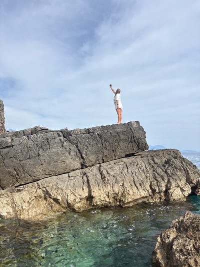 A woman is poised on a rocky cliff edge on a sunny day. She is wearing a white shirt and shorts, her body is parallel to the ground, arms extended. Below the cliff, clear water meets the rock face. The sky is blue with scattered clouds. The location is likely Seoca, Montenegro, inferred from the image's provenance and the terrain visible.