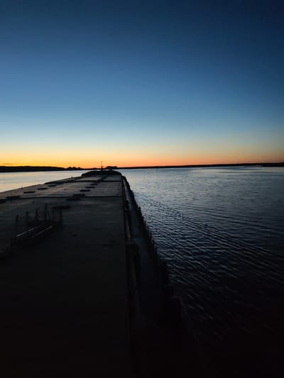 This is a vertical, medium-shot photograph taken at dusk on a concrete pier, likely in Irdomatka, Russia. The sky is a gradient of deep blue at the top, transitioning to a pale blue and then a bright orange and yellow band near the horizon, indicating the setting sun. The water is dark and choppy, with visible ripples and a wake trailing from the pier. The pier itself is a long, concrete structure extending into the water, casting a shadow to the left. On the pier, there are several large, rectangular concrete blocks, and a small, metal structure resembling a ladder or railing is visible on the left side. The far end of the pier has a raised section with what appears to be a light pole. The environment is serene and appears to be an outdoor recreational or industrial area near a body of water. No people are present in the image.