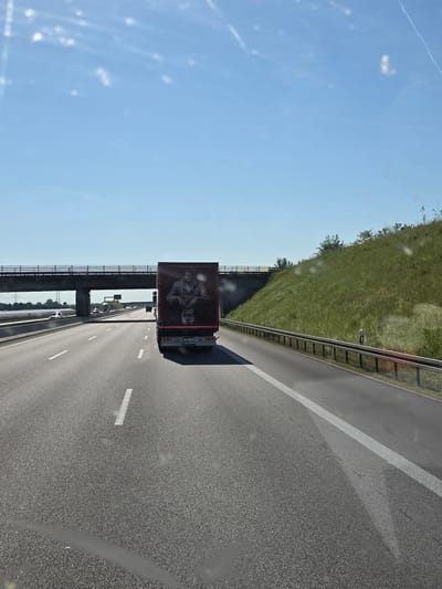 The image shows a view from inside a vehicle driving on a highway. The highway is paved with asphalt and marked with white lane dividers. To the right, a metal guardrail separates the highway from a grassy hill. A black semi-truck with a large black-and-white advertisement on its trailer is driving in the adjacent lane. The advertisement appears to feature portraits of two men. An overpass is visible ahead, spanning the highway. The sky is clear and blue. The location is likely on a highway in Barbing, Germany.