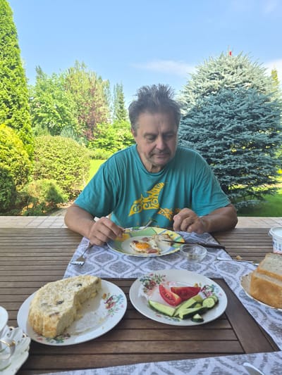 An outdoor portrait shows a middle-aged man with messy dark hair and a beard, wearing a teal t-shirt with a yellow graphic. He is seated at a wooden table, eating a meal. In front of him is a plate with fried eggs and ham, a fork, and a small glass bowl. To his left, on the table, are a plate with a slice of cake and a plate with sliced tomatoes and cucumbers. To his right, a white container and a plate with slices of bread are visible. The background features lush green trees and a bright blue sky. The man appears to be in a residential backyard, possibly in Vlast Truda, Russia, given the surrounding vegetation and outdoor setting.