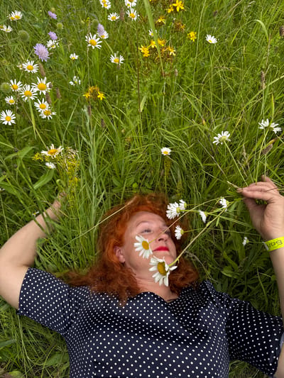 A woman with bright red hair lies in a grassy field, her head tilted back and eyes closed. She is wearing a dark blue dress with white polka dots and a yellow wristband with the word "БОЛ" printed on it. White daisies are scattered around her, with one resting on her cheek and another near her eye. Her left hand is raised, gently touching a few of the flowers, while her right arm rests at her side. The scene is set in a lush green field, with a variety of wildflowers and tall grasses. The lighting suggests it is daytime, possibly overcast, as there are no harsh shadows. The overall impression is one of peaceful repose and connection with nature. The presence of the wristband suggests she might be at an event or festival. The location is identified as Privolzhsky, Russia.