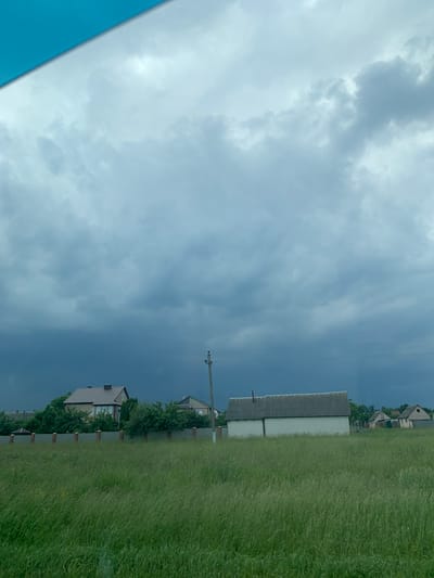 A wide, horizontal shot captures a rural landscape under a dramatic, cloudy sky. In the foreground, tall, green grass sways gently, characteristic of a field. A low, gray fence with intermittent brick posts runs across the mid-ground, separating the field from a row of houses and buildings.
To the left, a two-story house with a brown roof and cream-colored walls is partially obscured by lush green trees. To its right, a long, low building with a gray, corrugated roof and white walls stands prominently. Further to the right, a few smaller houses with pitched roofs are visible, nestled among trees and greenery. A lone utility pole with electrical wires stretches across the scene, adding a touch of man-made infrastructure to the natural environment. The sky above is a mix of dark, stormy gray clouds and lighter, wispy formations, suggesting an approaching storm or the aftermath of one. The overall impression is one of a peaceful, yet potentially unsettled, rural setting. The image is located in Слабада-Кучынскі сельскі Савет, Belarus.