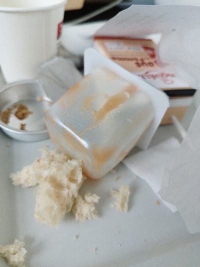 The image depicts a close-up view of discarded food items and packaging on a light-colored surface, presumably a table or tray, located in Clamart, France. In the immediate foreground and center, a translucent, rectangular plastic container lies on its side, smeared with a dried orange-brown residue, indicating it was a single-serving portion of a condiment or sauce. Adjacent to this container, to the lower left, is a cluster of torn, light-colored bread crumbs or pieces. To the left and slightly behind, a small, round, silver-colored metal dish contains a small amount of dark brown, granular residue. In the upper left background, a white disposable cup is partially visible. Towards the right background, crumpled white paper or napkin pieces are scattered. Further in the background, a cardboard food package, possibly from the "Bonne Maman" brand, is partially visible with red text. The scene presents a post-consumption state of a meal.