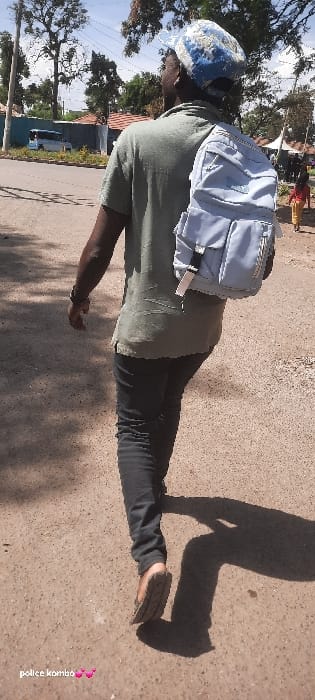 The image displays an outdoor street scene in Homa Bay, Kenya, under clear daylight. A dark-skinned individual, observed from behind, walks away from the camera, moving from the right side of the frame towards the left. This person wears a blue and white patterned baseball cap, a light olive green short-sleeved collared shirt, dark grey or black jeans, and brown open-toed sandals. A light blue backpack with multiple compartments and a white tag is worn on their right shoulder. A dark watch is visible on their left wrist.
The ground is an unpaved, light brown surface, likely dirt or gravel, showing faint tire tracks. A prominent, elongated shadow of the walking individual is cast on the ground to their right.
In the middle ground on the left, a paved road with a white line is visible, beyond which a light blue minibus is partially obscured by foliage and a fence. Further back, buildings with reddish-brown tiled roofs are present. Tall trees with dense green foliage line the road, casting dappled shadows.
On the right side of the frame, a small child, wearing a red top and light-colored trousers, walks further down the path in a similar direction to the main subject. Behind the child, white, tent-like structures, possibly market stalls, are visible. The sky above is a clear blue with minimal cloud cover. An overlaid text annotation, "police kombo ๐๐", is present in the lower-left corner of the image.