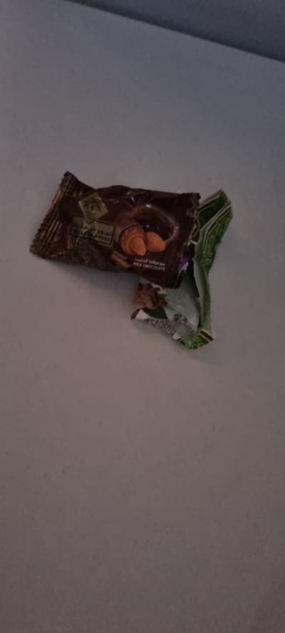 A crumpled milk chocolate candy wrapper lies on a plain, lightly textured surface. The wrapper is predominantly dark brown with some green and white accents. The front of the wrapper features an image of three almonds in milk chocolate. Text on the wrapper is visible in what appears to be Arabic and English. The English text reads "MILK CHOCOLATE" and "DATES". A green and white patterned wrapper is partially visible, folded over the chocolate wrapper. The scene is dimly lit, suggesting an indoor setting with artificial lighting. There are no people or animals visible. The overall impression is that of discarded packaging. The location context provided is Обшорон, Tajikistan.
