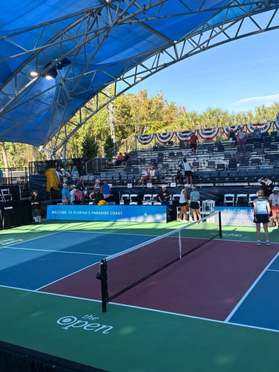 This image depicts an outdoor pickleball event under a clear, bright blue sky on a sunny day. The focal point is a pickleball court, distinguished by its blue, maroon, and green playing surfaces, separated by white lines. A net divides the court in the foreground.
To the right of the court, tiered bleachers are partially filled with spectators observing the game. These bleachers are decorated with red, white, and blue American flag-themed bunting. People of various ages are casually dressed, sitting or standing, watching the activity on the court.
Along the court's edge, a blue banner prominently displays the text "WELCOME TO FLORIDA'S PARADISE COAST." On the green area of the court, the white lettering "the Open" is visible, featuring a stylized pickleball logo.
To the left, a large, blue-fabric covered structure supported by metal scaffolding provides shade, under which additional people are gathered. Green trees and palm trees are visible in the background, suggesting a tropical or subtropical outdoor location, consistent with "Florida's Paradise Coast" and the provided Naples, United States context. The scene conveys an atmosphere of leisure and sports enjoyment during daylight hours.