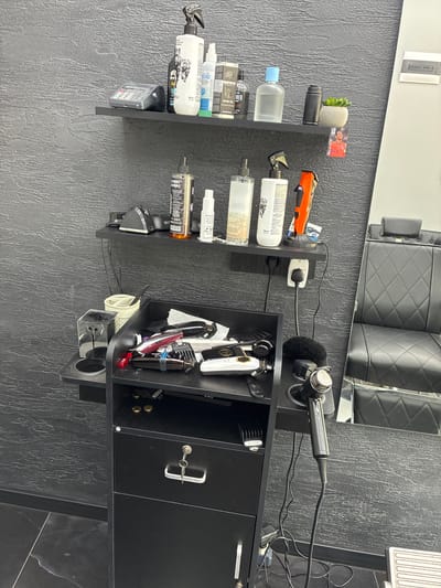 The image displays a well-organized section of a barber shop or hair salon, set against a dark gray, textured wall. A large mirror reflects a black, quilted barber chair in the background, with a dark, possibly tiled floor visible in the lower right. The overall location context is Simitli, Bulgaria.
On the left side, a vertical black shelving unit holds various grooming products and accessories. Items include a small artificial succulent plant, a card displaying a man's face and partial text "VERA VAN DE... PERE," and several bottles. These bottles appear to contain different liquids, with visible labels such as "BEARD OIL," "500 ml," "EMID," "BARBER," and partially seen "PURE" and "DOUBLE." A small calculator is also placed on one of these shelves.
To the right, a black, multi-compartment mobile station or trolley is featured. A power strip is visible on its upper surface, providing power to a professional tool, possibly a hair dryer or finishing machine, labeled "GAME PRO," which rests in a dedicated holder. The station's open compartments are filled with an array of hair styling tools: several hair clippers/trimmers (one red with a "SIC" logo), various clipper guards, scissors, and a comb. A small red clip also bears the "SIC" logo. A closed cabinet section of the station has a key inserted into its lock. More bottles and spray products are stored on a lower shelf of this station.
The scene, devoid of people, suggests a professional grooming workspace ready for use, with all tools and products neatly arranged and accessible. The lighting is bright, indicating an indoor setting.