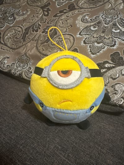 This is a close-up, overhead shot of a yellow plush Minion toy, likely a souvenir or collectible. The Minion is positioned on a textured surface, possibly a couch or armchair, which is upholstered in a dark, coarse fabric. To the left of the Minion, a portion of a patterned textile is visible. This fabric features an intricate, repeating design in shades of brown and gray, with paisley-like motifs and floral elements.
The Minion itself is round and plump, with its characteristic single eye covered by a silver goggle. The goggle is detailed with a black band and a white lens, revealing a brown iris and pupil. The body of the Minion is primarily yellow, with a light blue jumpsuit-like covering around its back and sides. Yellow stitching details and what appear to be buttons are visible on the blue fabric. A thin, bright yellow cord or loop protrudes from the top of the Minion's head. The toy appears to be in good condition, with no visible damage.
The lighting in the image is somewhat dim, suggesting an indoor setting, possibly in the evening or on a cloudy day. There are no clear indicators of the time of day, weather, or specific location beyond the provided context of "Змейка, Russia." The scene is static, with the toy simply resting on the surface. There is no text visible in the image.
