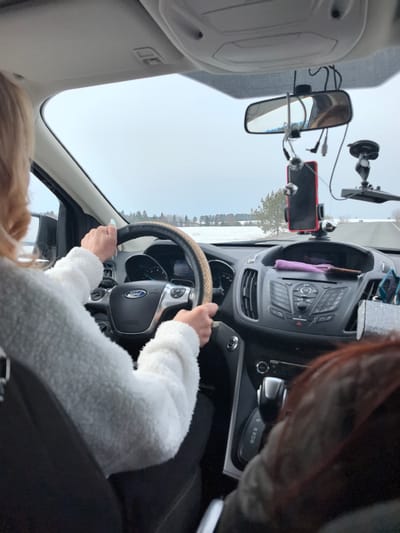 The image captures the interior of a vehicle, specifically a Ford, from a rear-passenger viewpoint, looking forward through the windshield. The scene is set in Kvarsa, Russia. A driver, identified by blonde hair and a white textured long-sleeved garment, is positioned in the left front seat, with both hands gripping the steering wheel, which is adorned with a light brown, woven decorative cover. The driver's fingernails are painted light blue. The dark dashboard displays vehicle controls, with a purple fabric item and a dark cylindrical object resting on its surface. A red smartphone is affixed within a suction-cup mount below the rearview mirror, which exhibits a visible crack and has several cables connected to it. The back of a passenger's head, featuring dark reddish-brown hair, is partially visible in the front right seat. Beyond the windshield, an expansive, snow-covered landscape with scattered evergreen trees stretches towards a distant horizon beneath an overcast sky, indicating an open road or rural setting.