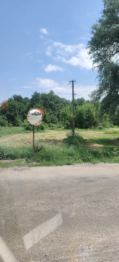 A convex traffic mirror on a metal pole stands next to a dirt road in Kolomak, Ukraine. The mirror reflects a cloudy blue sky and a dense forest of green trees. A wooden utility pole with wires runs parallel to the road, and a thick line of grass and wildflowers borders the roadside. The foreground shows the rough texture of the dirt road, with a hint of a white line painted on it. The scene is bathed in bright daylight under a clear sky with scattered clouds.