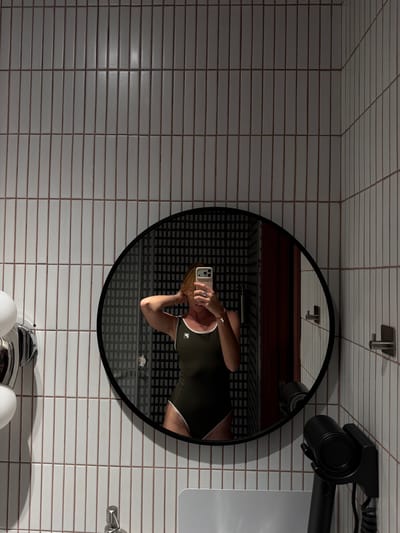 A person is reflected in a round black-framed mirror, standing in a bathroom with white tiled walls. The person is wearing a dark green swimsuit with white trim and a white logo. They are holding a smartphone in their right hand, taking the photo, and their left hand is near their head. The mirror reflects a patterned black wall behind the person. To the left of the mirror, a chrome fixture with white spherical components is visible, likely a light or soap dispenser. To the right of the mirror, a black hairdryer is mounted on the wall. A faucet is visible in the lower left corner, and the edge of a white sink or countertop is in the lower part of the image. The scene is indoors, in what appears to be a hotel bathroom. The image does not provide any specific information that would indicate a location in Kaji-Say, Kyrgyzstan.