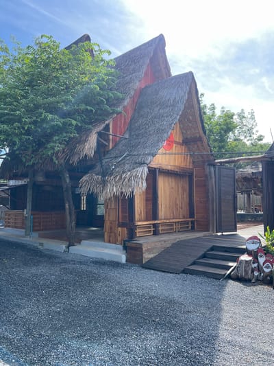 This media file depicts a rustic, open-air building in Tambon Pa Kae Bo Hin, Thailand. The structure features a traditional thatched roof made of dried palm leaves, supported by wooden beams. The walls are constructed from rough-hewn wooden planks, giving it a natural and slightly weathered appearance. In front of the building, there is a pathway covered with dark gray gravel.
To the right of the building, a wooden staircase leads up to a raised platform or deck. On this platform, there are decorative objects made from logs, painted to resemble figures, possibly gnomes or Santas, with red hats and white beards. A red circular sign with white text is visible on the side of the building.
Lush green foliage, likely trees and vines, surrounds the building, with some branches extending over and around the thatched roof. The sky above is partly cloudy, suggesting it is daytime. The scene conveys a sense of tranquility and a connection with nature.
The visible text on the red sign appears to be part of a name, possibly including the words "the orange cafe."