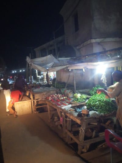 The image depicts a nighttime scene at an outdoor market, likely in the city of Baccure Kofare, Nigeria. Several individuals are present at various market stalls. In the foreground, a person wearing a red shirt and orange pants is crouching on a wooden crate. To the left, a person in dark clothing and white pants stands near a white vehicle. Further back, a stall displays piles of produce, including green leafy vegetables and red onions. A bright light illuminates the market area, casting shadows. The background shows a building structure and other market activity.
