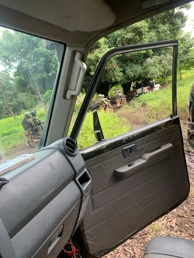 The image depicts a rural or semi-rural scene in Mkavior, Nigeria, viewed from the interior of a vehicle, likely an SUV or pickup truck. The front passenger door is open, revealing the grey interior door panel with its handle, window switch, and general wear. The dashboard, A-pillar, and a sun visor are also visible inside the vehicle, with "SRS AIRBAG" printed on the glove compartment area.

Outside the vehicle, a muddy, unpaved track extends into a densely vegetated area with numerous trees and green foliage under an overcast sky. On this track, two individuals wearing military-style camouflage uniforms, helmets, and tactical gear are riding a motorcycle away from the camera. The driver and passenger both have significant backpacks or equipment. Further down the track, beneath a large tree, a white Toyota pickup truck is visible, with multiple individuals (approximately 4-6) in its bed, also appearing to wear dark clothing or uniforms, some possibly armed with rifles. On the far right edge of the frame, a partial view of another person in a camouflage uniform is visible, standing outside the camera's vehicle. The overall scene suggests a patrol or movement of armed personnel in an off-road environment.