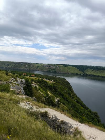 The image displays an elevated, panoramic view of a wide river valley in Horaivka, Ukraine, under an overcast sky. The foreground features a steep, grassy hillside characterized by exposed light-colored soil and rocks, with a faint dirt path traversing diagonally from the lower left. The midground is occupied by a broad, dark blue-green river flowing horizontally across the frame. The river is bordered on the near side by a densely vegetated, steep bank, which descends into the water. On the opposite bank, a continuous line of prominent layered rocky cliffs and lush green hills stretches across the scene. Several small, light-roofed structures are discernable amidst trees on the far bank, near the water's edge. A group of approximately 10-15 light-colored livestock, consistent with sheep or goats, is visible on the near bank's slope, positioned closer to the river. The sky overhead is largely covered by various formations of grey clouds, with a small patch of lighter blue sky visible towards the upper left horizon. No human subjects are present. The scene depicts a natural, rural environment.