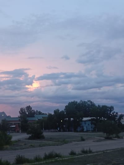 The image depicts an overcast sky with hints of pink and yellow near the horizon, suggesting either sunrise or sunset. A rural or suburban landscape unfolds below, characterized by a dirt road or unpaved area in the foreground. Beyond this, several low-rise buildings are visible, interspersed with trees. One building appears to be brick with a green roof, while another is a lighter color, possibly blue. The overall impression is one of a quiet, perhaps sparsely populated area, possibly in the city of Shagonar, Russia, given the context. No people or animals are discernible in the scene. The lighting is dim, consistent with twilight conditions.
