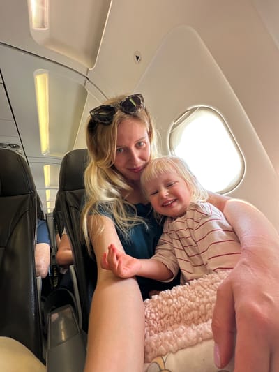 In the cabin of an airplane, a blonde woman with sunglasses on her head and a blue shirt smiles at the camera while holding a young blonde child in her lap. The child, wearing a striped shirt and a pink fuzzy blanket, grins widely. The airplane cabin has black leather seats and overhead lights. The image is cropped to focus on the woman and child, with the window and other passengers visible in the background. This image does not contain any elements that would indicate the location in Yehud-Monosson, Israel.
