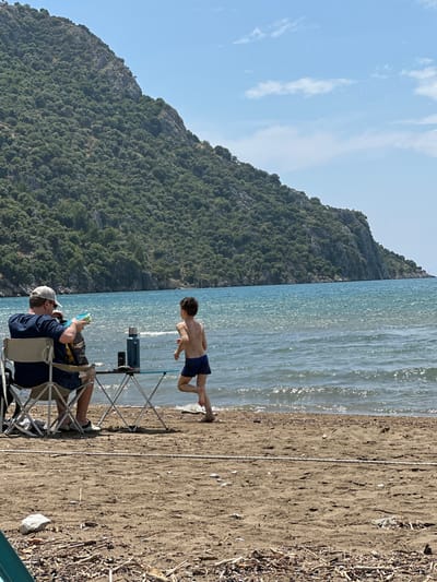 A beach scene in Ortaca, Turkey, shows a man seated in a folding chair on the sand, wearing a dark blue shirt, shorts, a white hat, and sunglasses. He is holding a blue object. A folding table stands nearby with a thermos and other items on top. A young boy in dark blue swim trunks is leaping towards the water's edge. The sea is sparkling in the sunlight. A large, green, tree-covered mountain is in the background under a blue sky with white clouds. Sticks and debris are scattered on the sand near the edge of the photo.