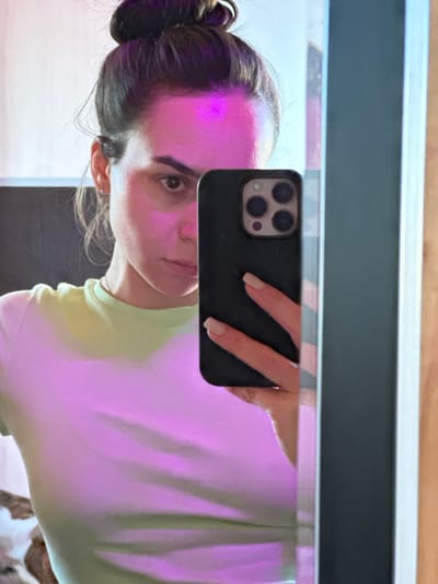 A female subject is depicted in a mirror selfie taken in an indoor setting within the city of 3-Y Uchastok, Russia. The individual, an adult with brown hair tied in a bun, has visible brown eyes and wears a light green short-sleeved top. Her right hand, with light-colored manicured fingernails, holds a dark-colored smartphone with a triple camera array, positioning it to capture her reflection. A distinct pink-purple light source illuminates the subject's forehead and upper torso. The environment is minimally visible, showing a dark vertical frame or object reflected on the right edge of the mirror, and a lighter, possibly wooden surface further to the right. A darker, undefined background element is reflected behind the subject's left shoulder. The subject's gaze is directed towards the smartphone camera.