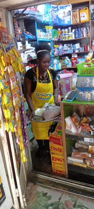 A woman wearing a yellow "ONGA" apron holds a tray with bags of goods inside a store. Shelves are stocked with various products, including "SuperFON", "Value Peak", and "KING'S" vegetable oil in "6x2L" cartons. Bags of "Chapa Chips" are displayed on the left. The image shows a typical small retail business setting.