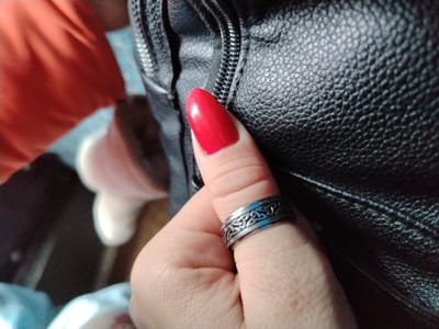 A thumb with a ring is seen in the foreground. The ring is silver with an intricate, dark pattern. The thumb has a red, almond-shaped nail. A black, textured, possible leather surface is behind the thumb and to the right. An arm with an orange garment is partially visible in the upper-left. A white or light-colored object appears blurred and out of focus in the background to the left. The bottom of the frame shows partially visible fabric.