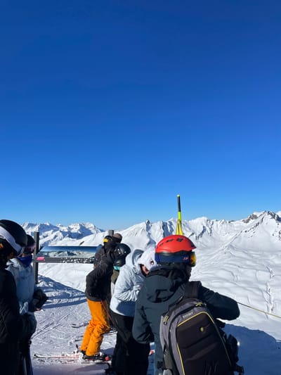 The image captures a vibrant winter sports scene high in the snow-covered mountains of Séez, France, under a brilliant, cloudless blue sky. The scene is bathed in bright sunlight, indicating excellent weather conditions for skiing or snowboarding.
In the mid-ground, a group of skiers and snowboarders is gathered on a snowy slope or platform, looking towards the expansive mountain range. They are all dressed in full winter sports attire, including helmets, goggles, and insulated jackets and pants.
Prominently featured is a person on the left wearing a bright red helmet with goggles, a grey ski jacket, and a black backpack displaying the "K2" logo. Adjacent to them is another individual in a white jacket and dark helmet. Further to the right, a person in a black jacket and distinctive bright orange ski pants stands on skis, which are partially visible and show the brand "NORDICA." Other individuals in dark ski gear are partially visible in the foreground, suggesting a larger group.
A yellow and black striped pole or marker stands upright in the snow behind the central group. A metal structure, possibly part of a railing, signpost, or terrain park feature, is also visible behind the people, with the word "SKATEBOARD" clearly printed on it. The background features endless snow-capped peaks and valleys, emphasizing the high-altitude, alpine environment. The clear visibility and bright conditions suggest a perfect day for mountain activities.