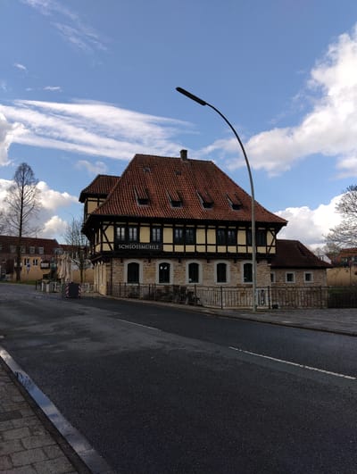 This image captures a daytime scene in Burgsteinfurt, Germany, featuring the Schlossmühle building.
The central subject is a two-story structure identified by the signage "SCHLOSSMÜHLE" on its facade. The building exhibits a traditional German architectural style with a stone foundation on the ground floor and a half-timbered upper story painted yellow with dark brown timbers. It features a prominent, steeply pitched roof covered in red clay tiles, interspersed with several small dormer windows. The windows on both floors are dark-framed and rectangular. A dark, curved street lamp post extends from the right foreground, curving over the road towards the building.
In the foreground, a dark asphalt road, slightly damp on its left edge, occupies the lower portion of the frame. A segmented white line is visible on the road surface. A stone-paved sidewalk runs along the left side of the road, and a metal railing borders the road adjacent to the Schlossmühle building. Behind this railing, several white, covered outdoor umbrellas and their bases are visible, along with a red and white striped barrier block, suggesting outdoor seating or an ongoing setup.
To the left of the Schlossmühle, further down the street, another building with a similar red-tiled roof and lighter facade is partially visible. A tall, bare deciduous tree stands prominently to the left of the frame, with other smaller bare trees scattered in the background. The sky is partly cloudy, displaying significant patches of blue with white stratocumulus and cirrus clouds. No individuals or vehicles are present in the immediate scene.