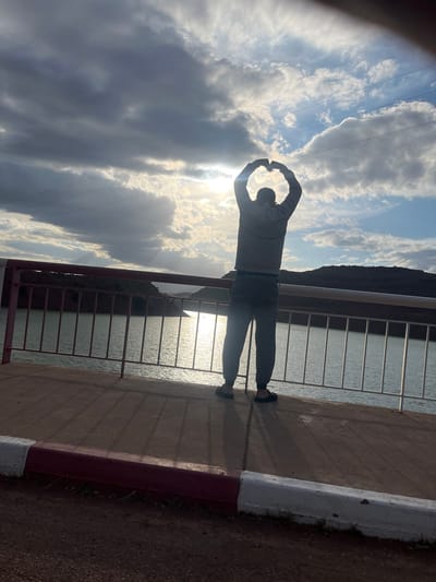 An eye-level outdoor photograph shows a person standing on a pier at Tabaroucht, Morocco with the ocean on one side of them, and the sky above them. The pier is made of light brown concrete and lined with a white and maroon barrier. A metal safety rail is attached to the edge of the pier. The ocean water sparkles due to reflected sunlight. The person faces away from the camera and is wearing gray pants and a gray hooded sweatshirt. The person's hands are raised and curved to form a heart-like shape with their fingers touching. The sky is cloudy, with some patches of blue visible through the clouds. Power lines are visible in the background of the image.