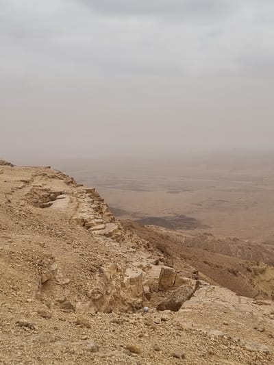 The image depicts the edge of a cliff overlooking a desert landscape. The cliff face is composed of layered rock and loose gravel. The landscape below appears hazy and indistinct, with some darker patches that may be depressions or water features. The location is likely Mitzpe Ramon, Israel, based on the desert environment and geological formations. There are no people or animals visible in the frame.