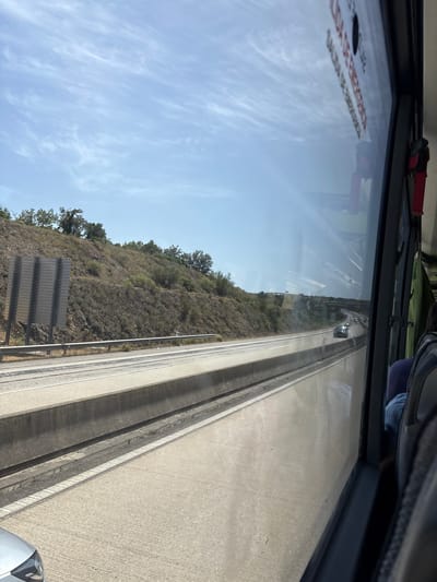 This is a medium shot, taken from inside a vehicle, looking out a window onto a highway in Albufeira, Portugal. The highway has multiple lanes, separated by concrete dividers. The terrain to the left of the highway is a dry, rocky hillside covered with scrub brush and a few scattered trees. A large, grey, slatted sign is visible on the hillside. Several cars are driving on the highway, and the sky is a clear blue with wispy white clouds. The window has reflections on it, including some text that appears to be warning signs. The right side of the image shows the interior of the vehicle, with seats and a window frame.
