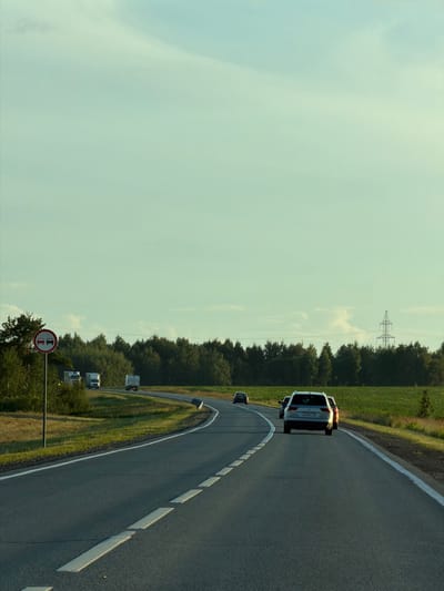 The image depicts a two-lane asphalt road curving through a rural landscape under a pale blue sky with scattered clouds. On the left side of the road, a circular traffic sign with a red border shows two red trucks, indicating a "no overtaking" zone for trucks. Several white trucks are visible on the road ahead, traveling in the same direction as the camera. A white SUV is in the foreground, also traveling on the road. The roadside is lined with green trees and grass, and a power line tower can be seen in the distance on the right. The scene appears to be set in the daytime, with the sun casting a warm light. The overall impression is of a typical highway scene in a wooded, rural area.