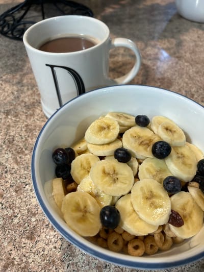 A close-up, eye-level forensic view captures a prepared breakfast or snack on a speckled countertop surface, likely granite, in Thousand Oaks, United States. In the foreground, a white ceramic bowl with a thin blue rim is filled with light brown, O-shaped cereal, generously topped with multiple circular slices of ripe banana and scattered dark blue blueberries. Some darker, reddish-brown dried fruit pieces are also visible among the cereal. To the left and slightly behind the bowl, a white ceramic mug with a prominent black capital letter "D" on its side contains a dark brown liquid, consistent with coffee or hot chocolate. The mug features a handle on its right side. A small portion of a black, ornate metal object, possibly a stand or decorative item, is visible in the upper left background, out of focus. No individuals are present in the scene.