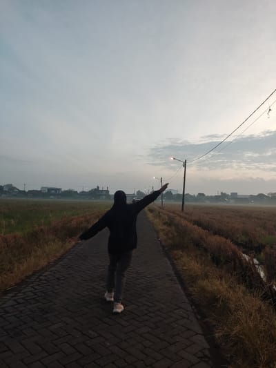 In Sukorejo, Indonesia, a person wearing a dark hooded jacket and grey pants walks along a paved road during dawn. The person, viewed from behind, has their arms outstretched, as if embracing the day. The road is bordered by dry, golden grass on either side, and in the distance, a low-lying town is visible under a hazy, pale blue sky with wispy clouds. Streetlights line the road, illuminating the path. Power lines crisscross the sky, adding a linear element to the scene. The overall atmosphere suggests a peaceful, early morning start to the day in a rural or semi-rural Indonesian setting.