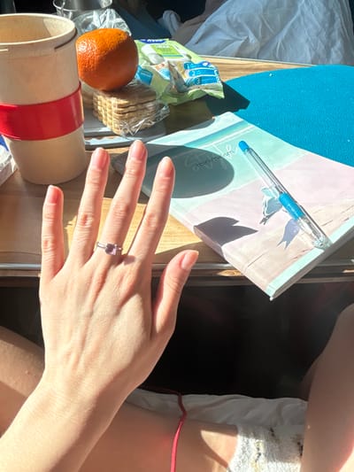 The scene depicts a close-up, high-angle view of items arranged on a wooden surface, identified as being in Kalachinsk, Russia. A fair-skinned left hand, adorned with a silver-colored ring featuring a square-cut purple gemstone on the ring finger, occupies the foreground. The hand exhibits manicured nails with a light, neutral polish.
On the left side of the wooden surface, a beige cylindrical cup with a red silicone band is visible. Adjacent to it, a single orange rests on a clear plastic-wrapped stack of approximately seven rectangular crackers. Behind these items, a partially opened green package with Cyrillic text, possibly containing wipes or snacks, is present.
To the right, a pastel-colored notebook or journal displaying "minimalism" on its cover lies on a light blue textured mat. A transparent blue ballpoint pen rests diagonally across the notebook. Strong natural light illuminates the scene, casting distinct shadows from the hand, pen, and objects. The lower portion of the frame shows the individual's lap, partially clad in light-colored fabric, with a red drawstring visible. A crumpled white fabric object is present in the background.