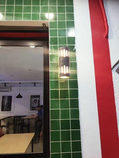 The image shows a close-up of a building exterior featuring a wall with green tiles, a red and white striped section, and a light fixture. In the reflection of a glass window, a person wearing a blue t-shirt and dark pants is visible, appearing to be seated at a table inside a cafe. The interior scene includes tables, chairs, and framed pictures on the wall. The building is located in Ariana, Tunisia.